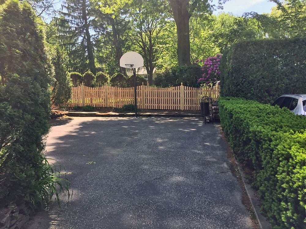 Asphalt driveway with wooden fence, basketball hoop, and green foliage.