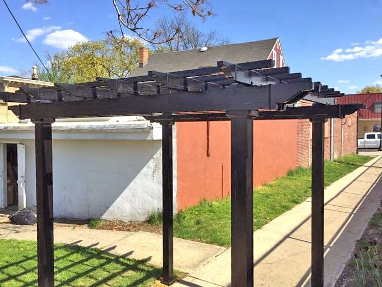 Black pergola in a yard with buildings and blue sky in the background.