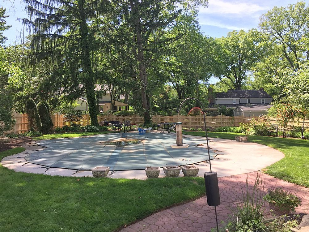 Backyard with a pool covered with a blue tarp. Green lawn and trees surround it.
