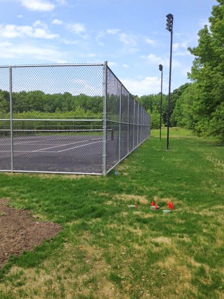 Tennis court enclosed by a chain link fence, with lighting poles in a grassy area, trees in the background.