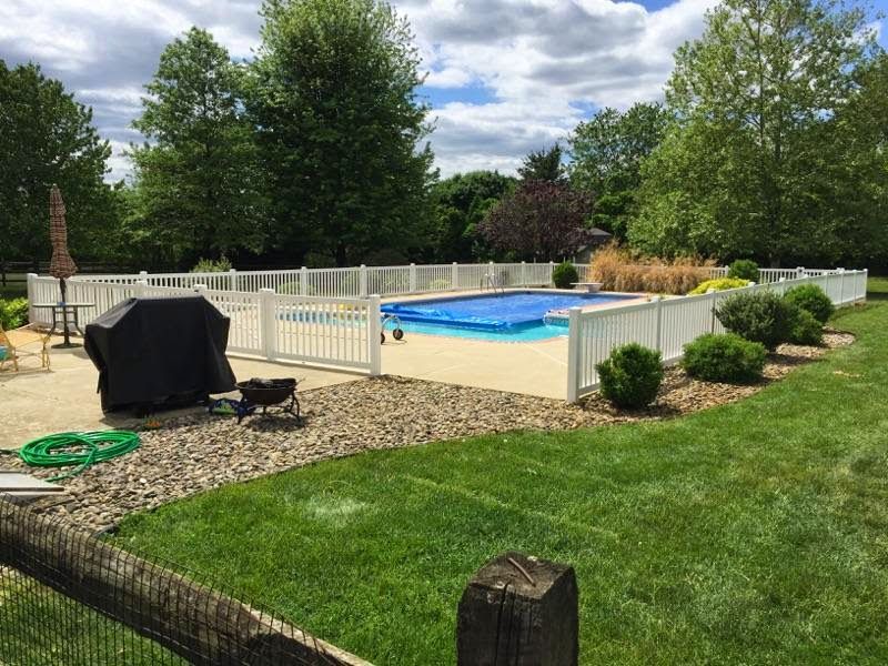 White fenced-in backyard pool with blue cover, grill, rock bed, and green lawn on a sunny day.