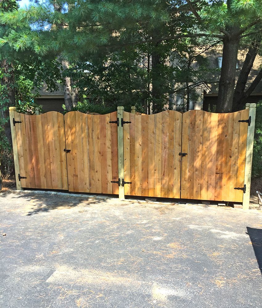 Wooden gate with scalloped top, black hinges, and posts, on a driveway.