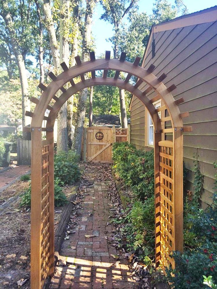 Wooden archway with brick path leading to a gate, flanked by plants and a house.