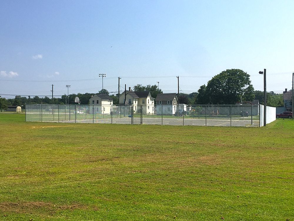 Open outdoor basketball court surrounded by chain-link fence on a grassy field; several buildings in background on a sunny day.
