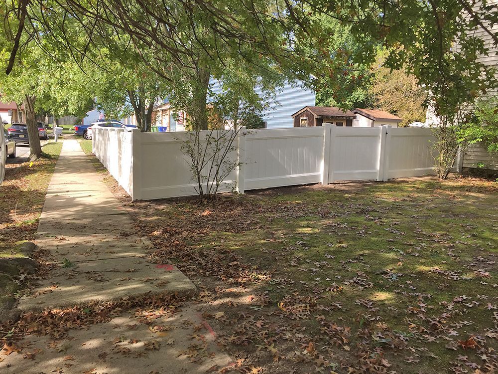 White fence bordering a sidewalk and yard, under trees with fallen leaves.