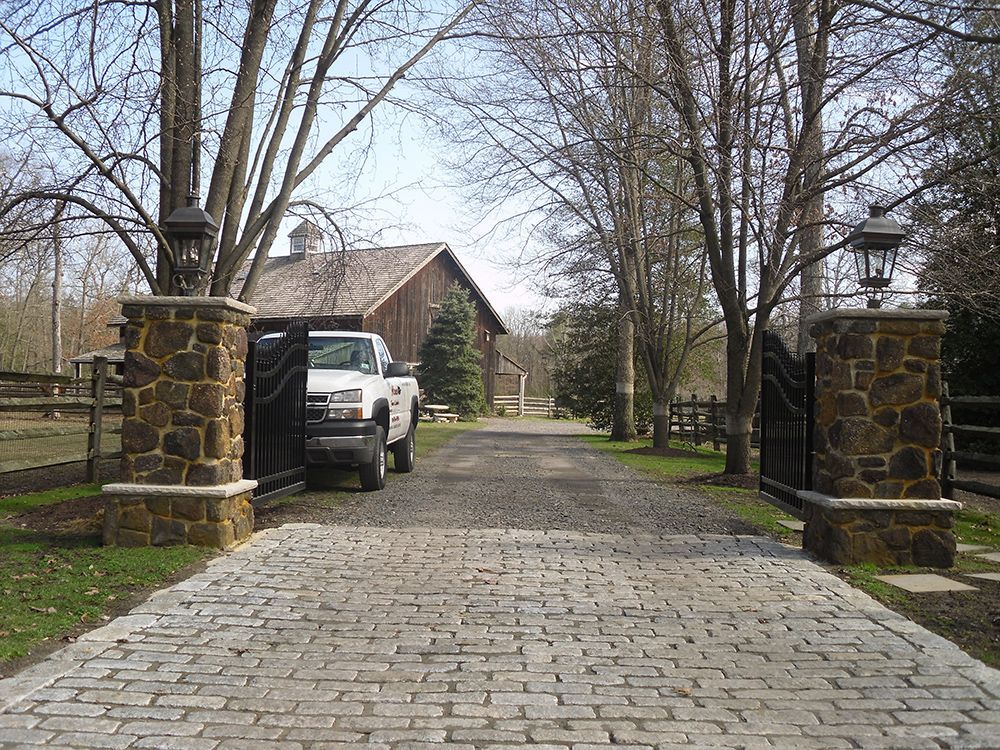 Stone pillars with ornate lamps and black gates open to a cobblestone driveway leading to a barn. A truck is parked.
