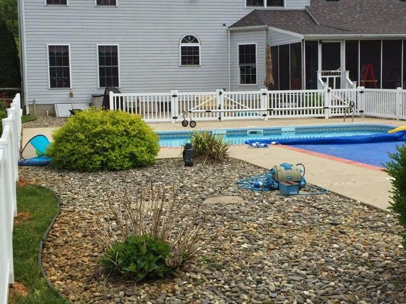 Backyard with a swimming pool surrounded by a white fence, gravel landscaping, and a house in the background.