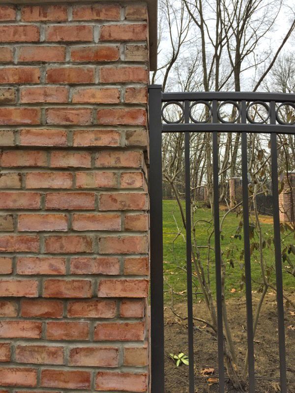 Red brick pillar next to a black metal fence with a grassy background and bare trees.