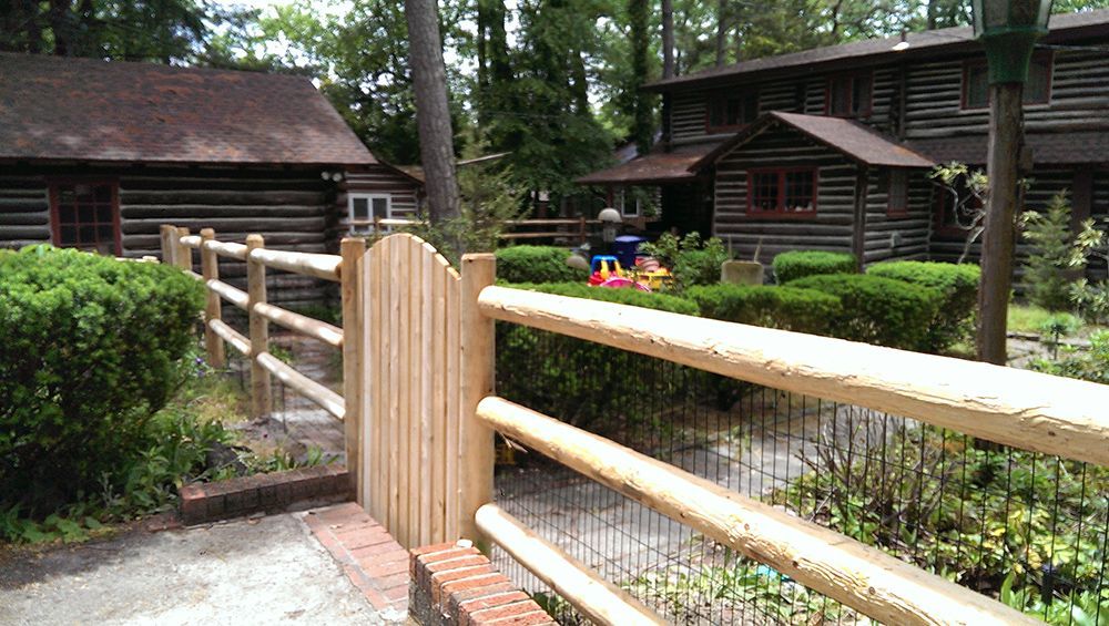 Wooden fence with gate leading to log cabins surrounded by greenery.
