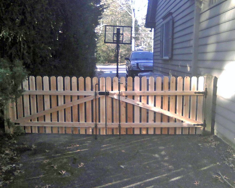 Wooden gate with vertical pickets, set in a yard, leading to a driveway with a basketball hoop.
