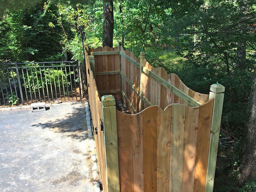 Wooden outdoor shower with scalloped fence, adjacent to trees and asphalt driveway.
