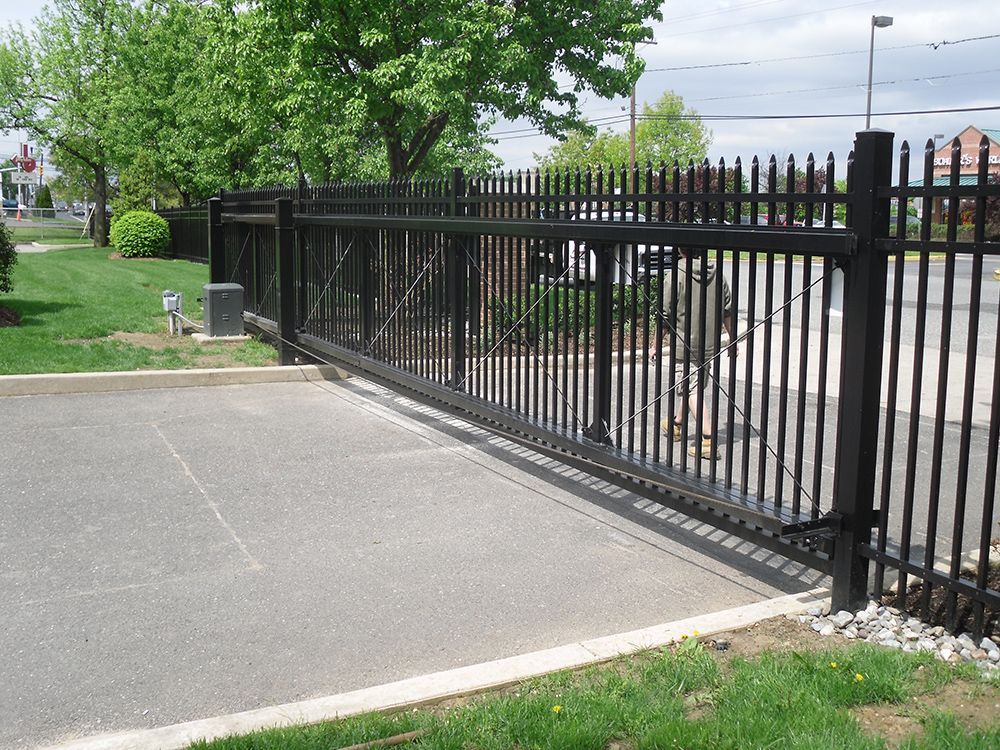 Black metal sliding gate in front of a parking area, green grass and trees visible.