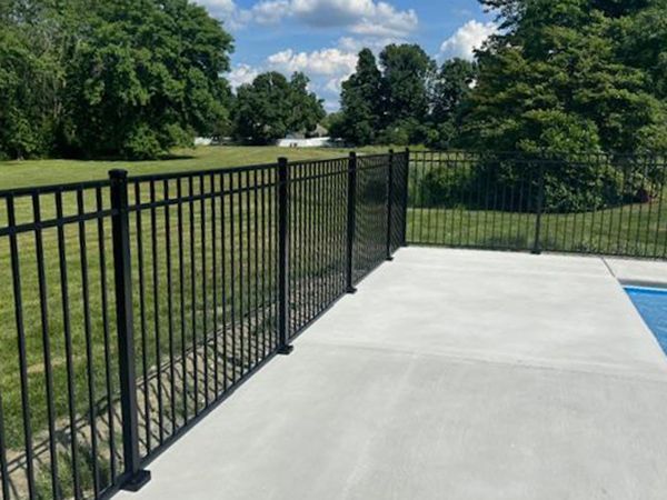 Black metal fence surrounding a concrete patio area next to a pool, with green grass and trees in the background.