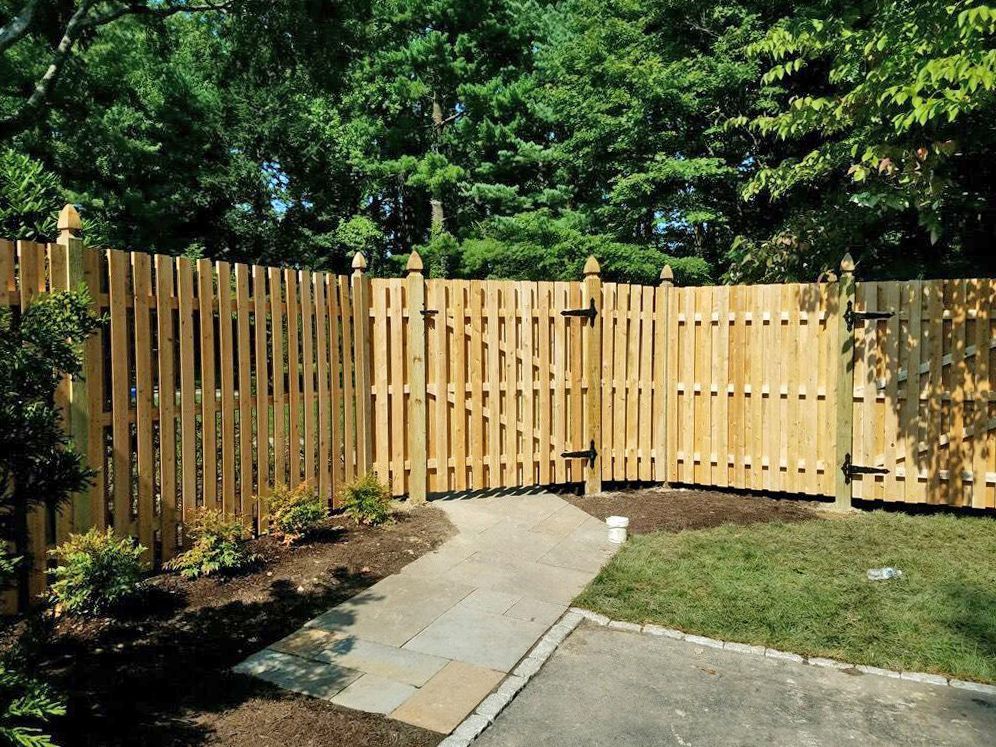 Wooden fence with gate, bordering a stone path and lush green grass. Trees in the background.