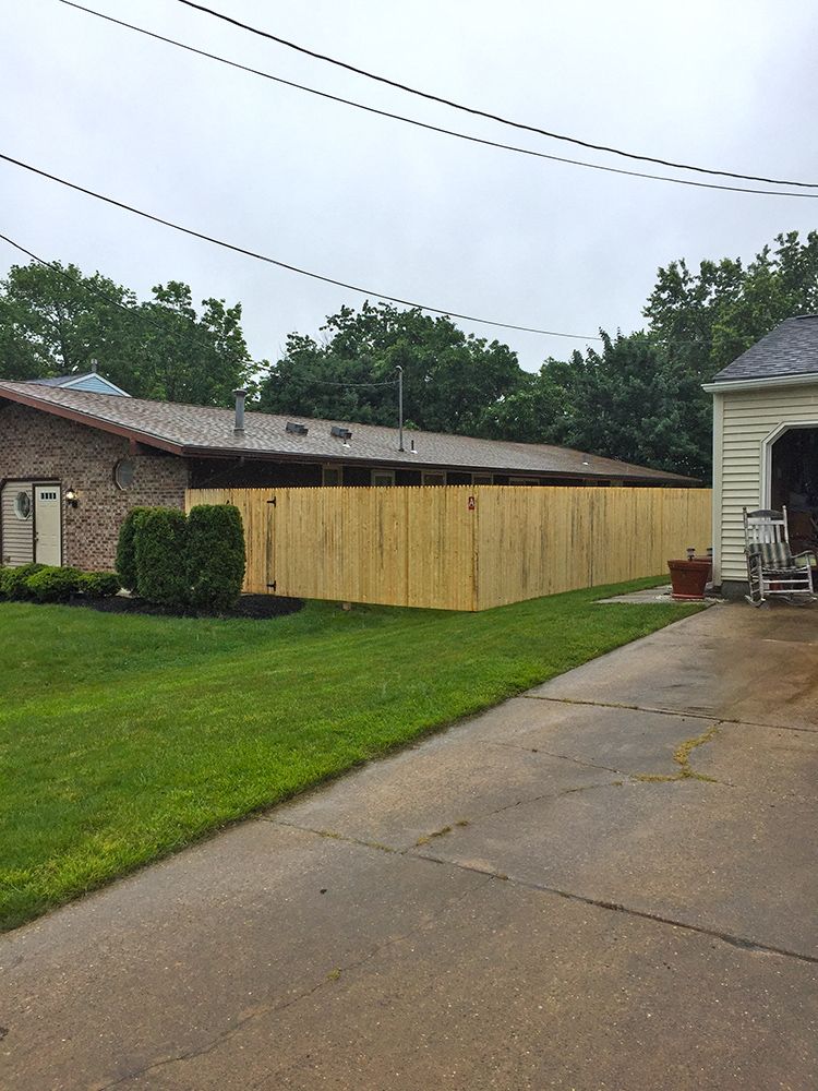 A newly constructed light-colored wooden fence runs along the side of a brick house, next to a driveway and lawn.