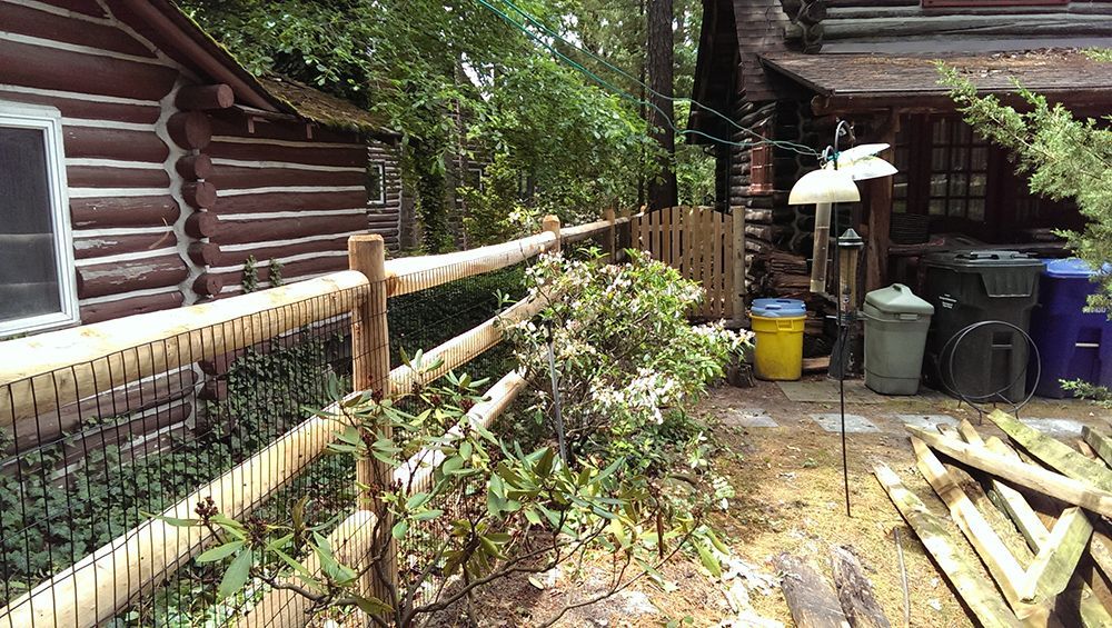 A wooden log cabin with a wooden fence. Bushes and trash cans sit nearby.