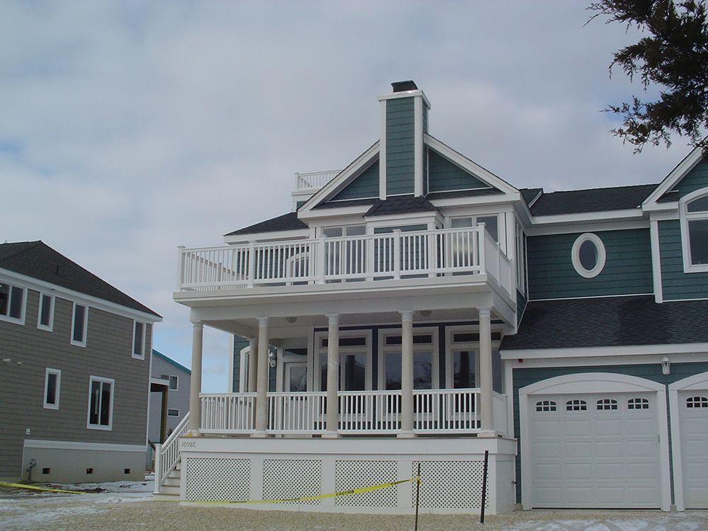 Two-story teal and white beach house with balcony and garage, under a cloudy sky.