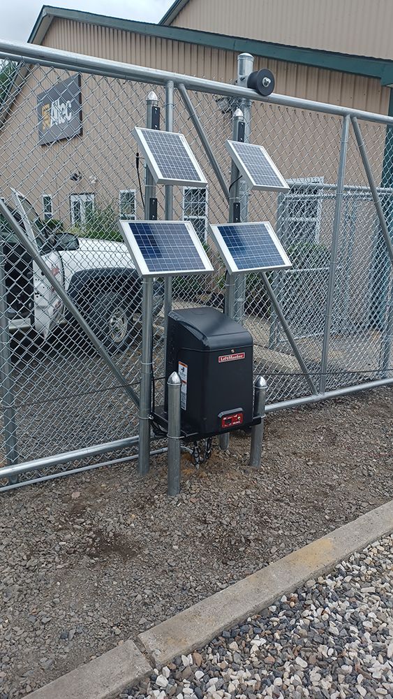 Solar panels powering an automatic gate opener on a chain-link fence, gravel ground, business in background.