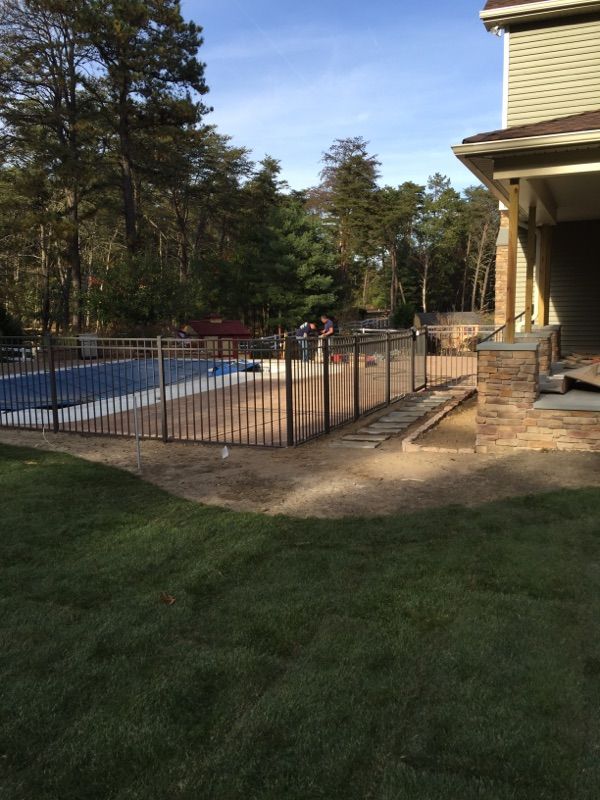 Brown metal fence surrounds a blue swimming pool in a backyard next to a house.