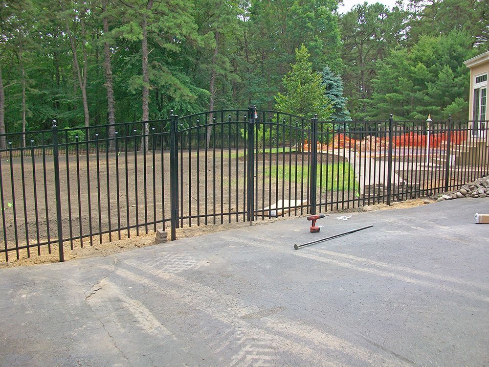 Black metal fence with gate, enclosing a yard with trees in the background. Asphalt driveway in the foreground.