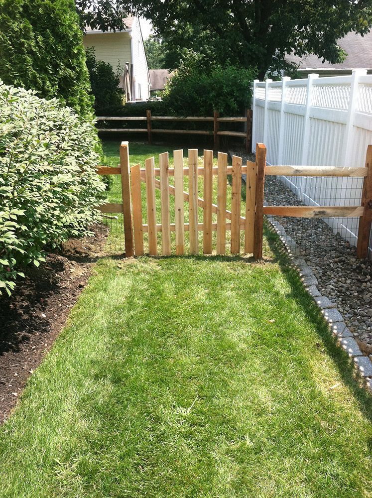 Wooden gate in a grassy yard, flanked by a split-rail fence and a white vinyl fence.