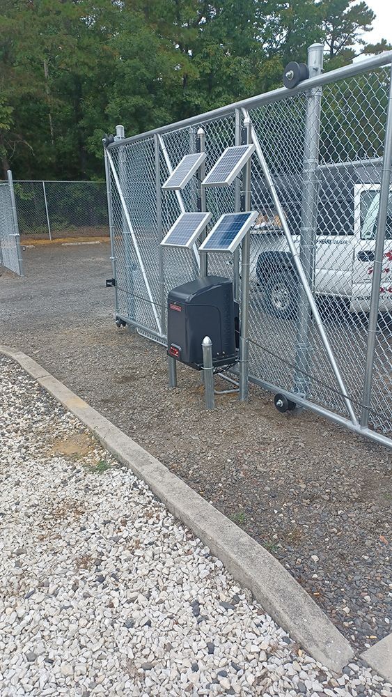 Chain-link gate with solar panels, a gate opener, and a white truck in a gravel area next to a curb.