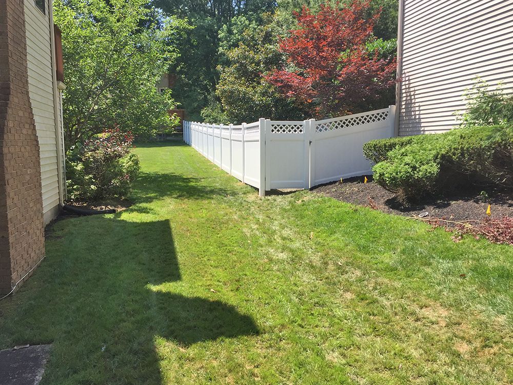Lush green lawn alongside a white picket fence, between two buildings, with shrubbery and trees in the background.