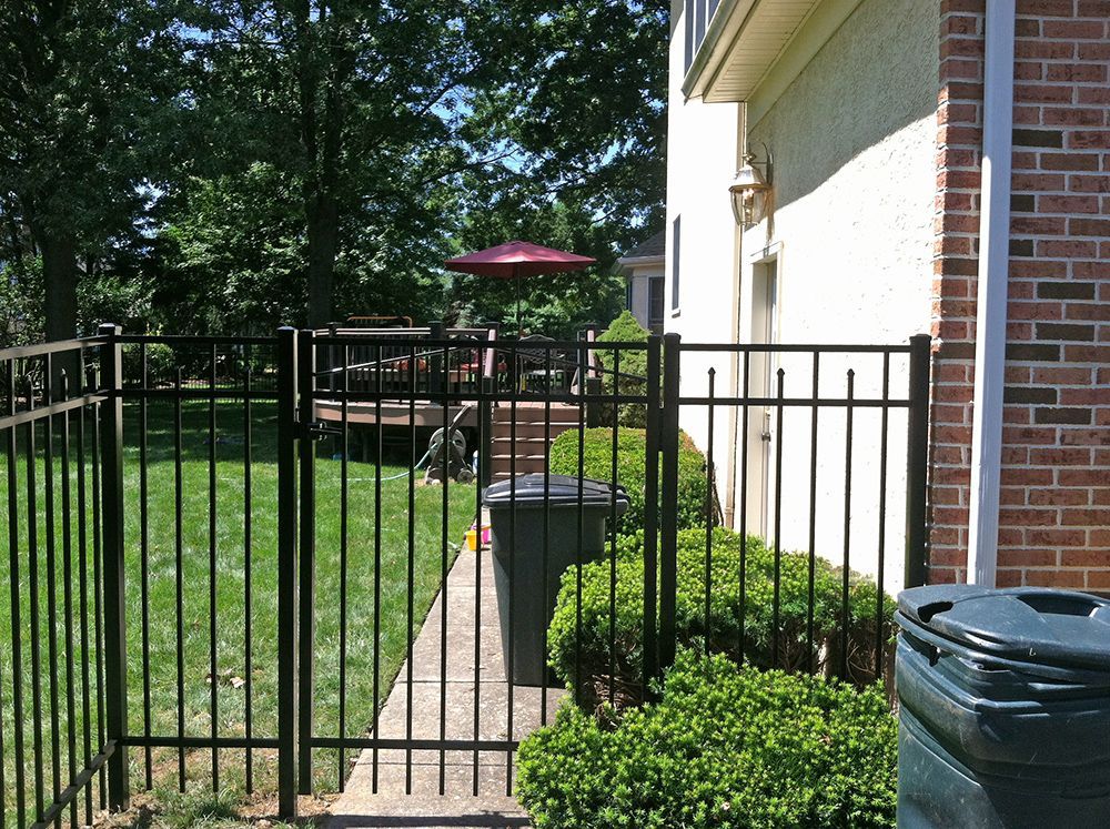 Black metal fence with gate, bordering a backyard with greenery and a house with a red brick facade.