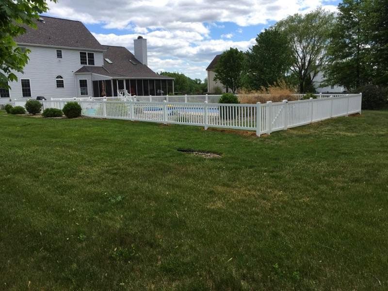 White fenced-in pool in a backyard, with a two-story white house and overcast sky in the background.