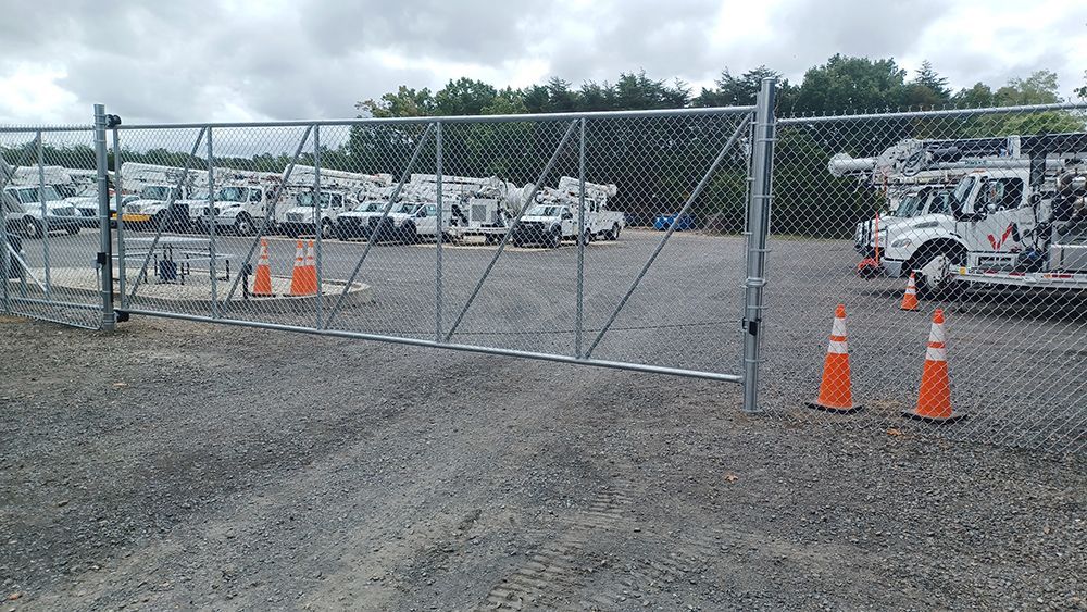 Chain-link fence encloses a gravel lot with utility trucks. Orange cones near the gate. Cloudy sky in the background.