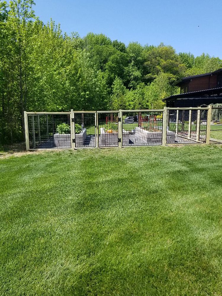 Garden bed enclosed by a wooden fence with gates, set in a grassy yard, with trees in the background under a blue sky.