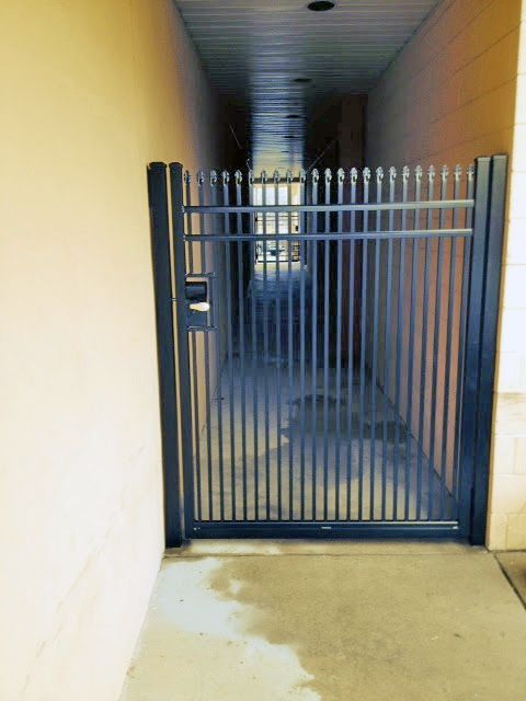 Black metal gate in a narrow passageway, flanked by beige walls and a concrete floor.