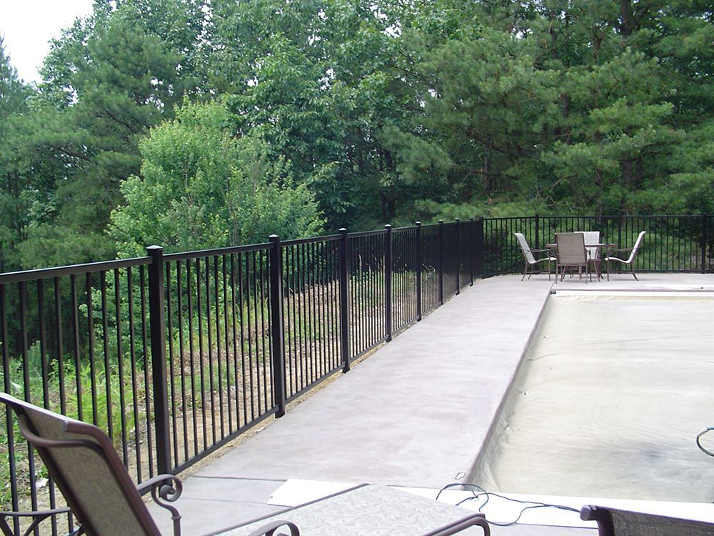 Black metal fence surrounds a concrete pool deck with chairs and a table, wooded background.