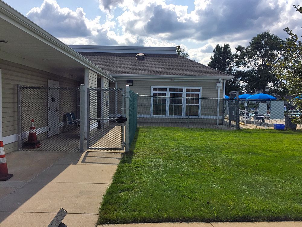 View of a building and a fenced-in lawn leading to a pool area with chairs and umbrellas under a cloudy sky.