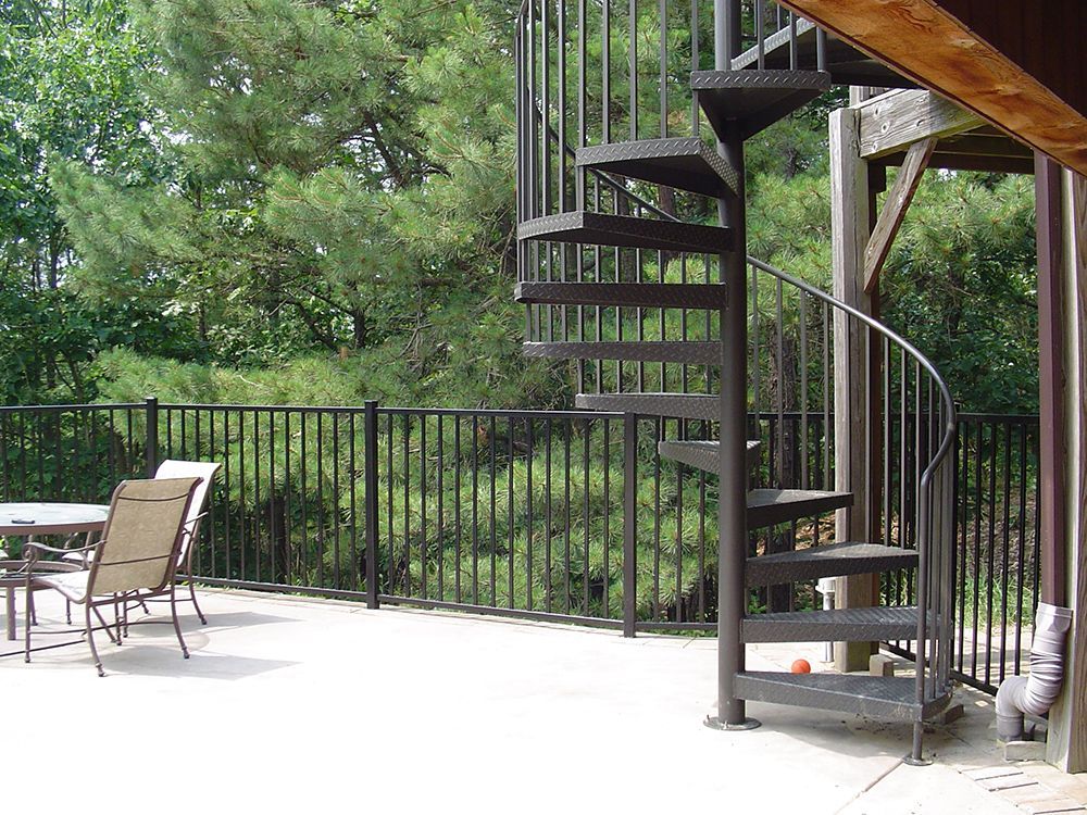Outdoor spiral staircase leading up to a deck with chairs and a table, black railing, trees in background.