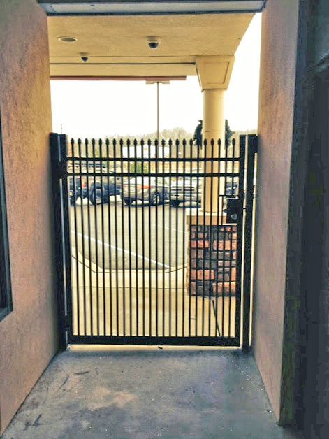 Black metal gate in a narrow passageway, leading to a parking area. Beige walls and a concrete floor.