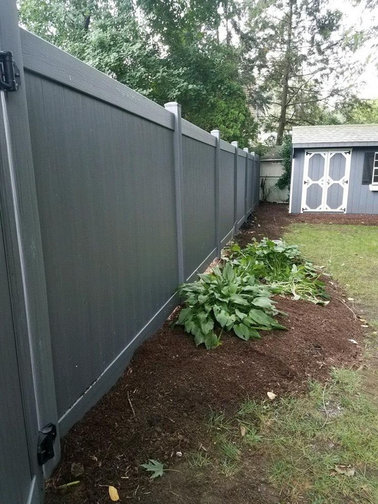 Gray vinyl fence borders a yard with mulch, plants, and a small shed.