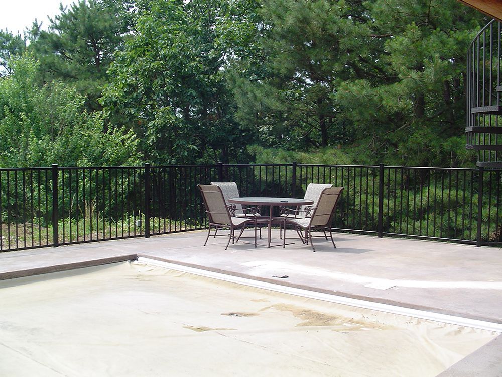 Patio with a table and chairs, surrounded by a black railing, and lush green trees in the background.