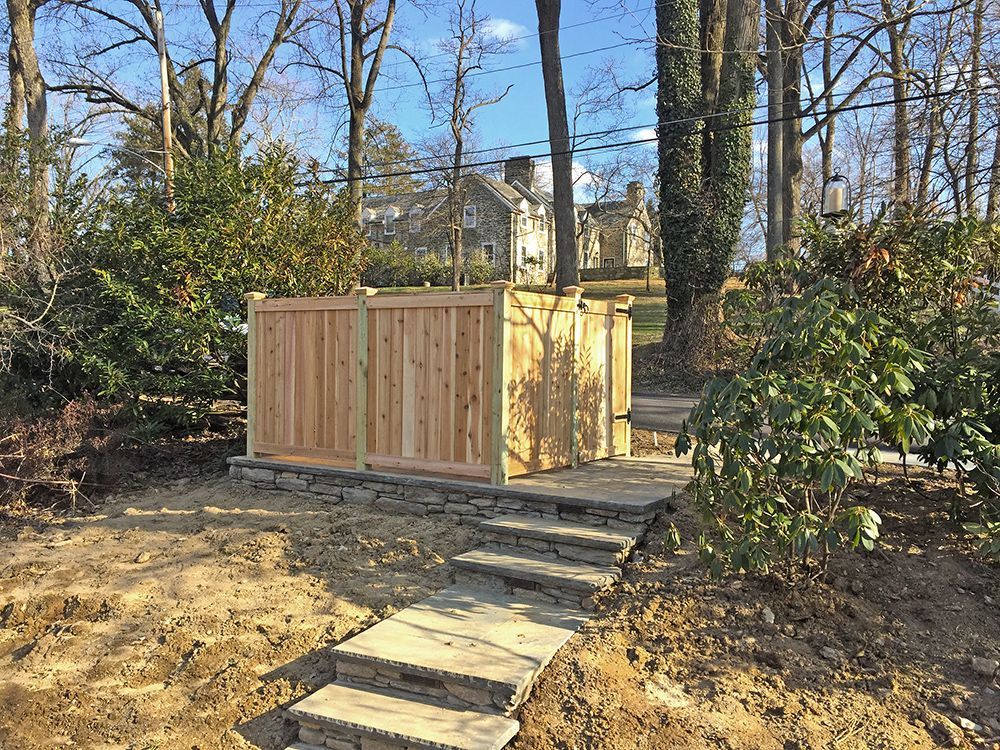 Stone steps lead up to a wooden privacy fence, surrounded by trees and shrubs in a yard.