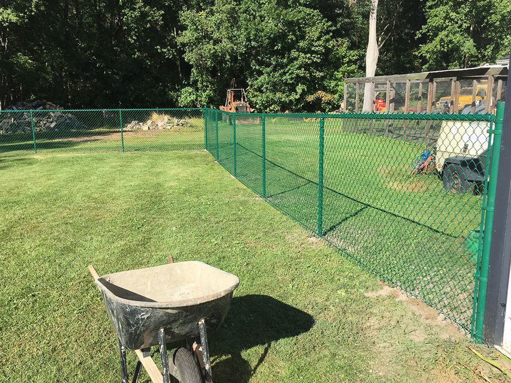 Green chain-link fence encloses a grassy yard; a wheelbarrow filled with cement sits in front.