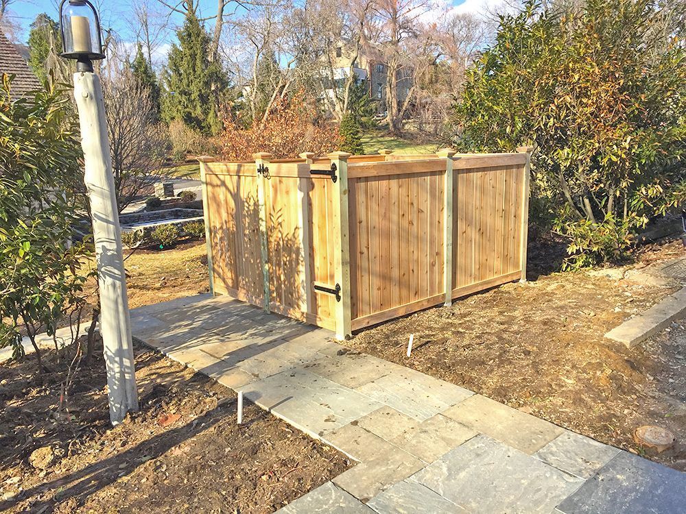 Wooden fence with gate, enclosing an area, next to a stone walkway and lamp post in a yard.