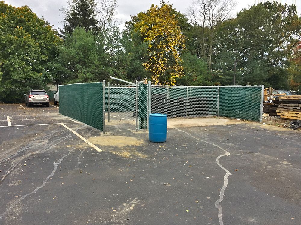 Chain link fence enclosure with green privacy screen in a parking lot. A blue barrel is inside.