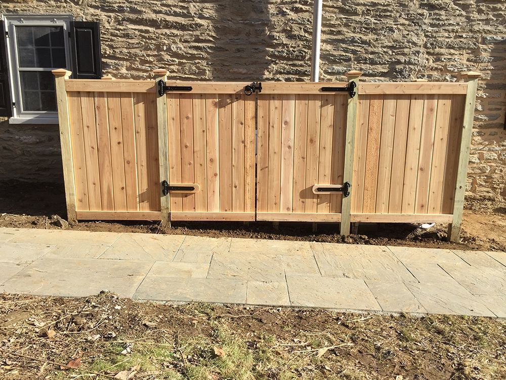 Wooden trash bin enclosure next to a stone building, with two hinged doors and a stone walkway in front.