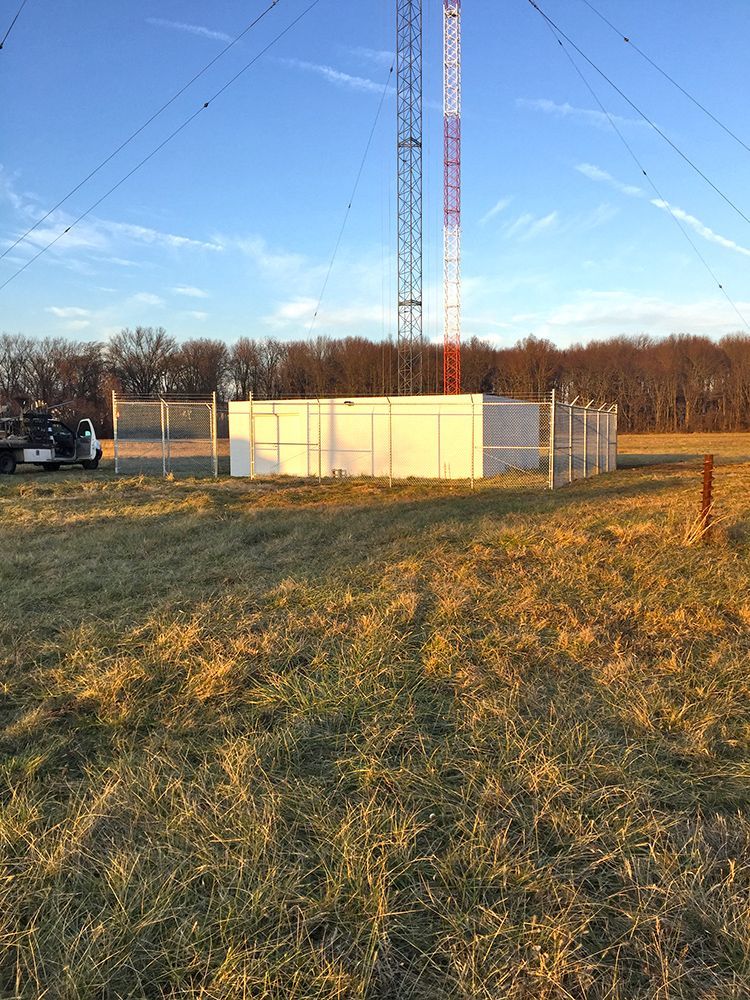 Two radio towers with support buildings fenced in a grassy field.