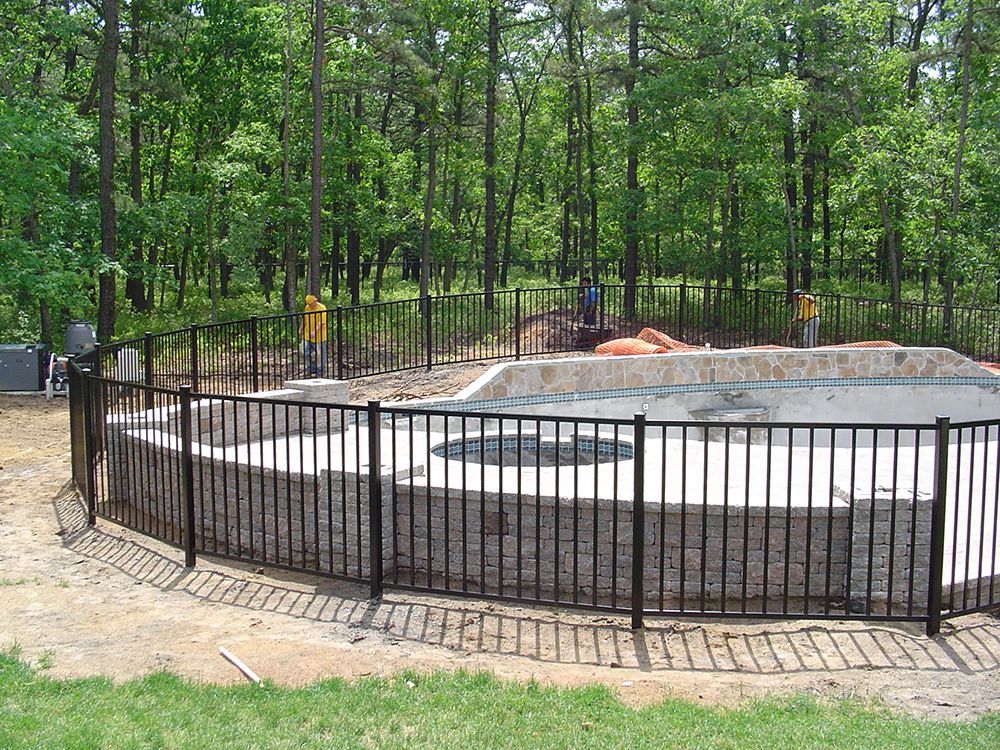 Black fenced-in, unfinished pool area with stone walls, surrounded by trees.