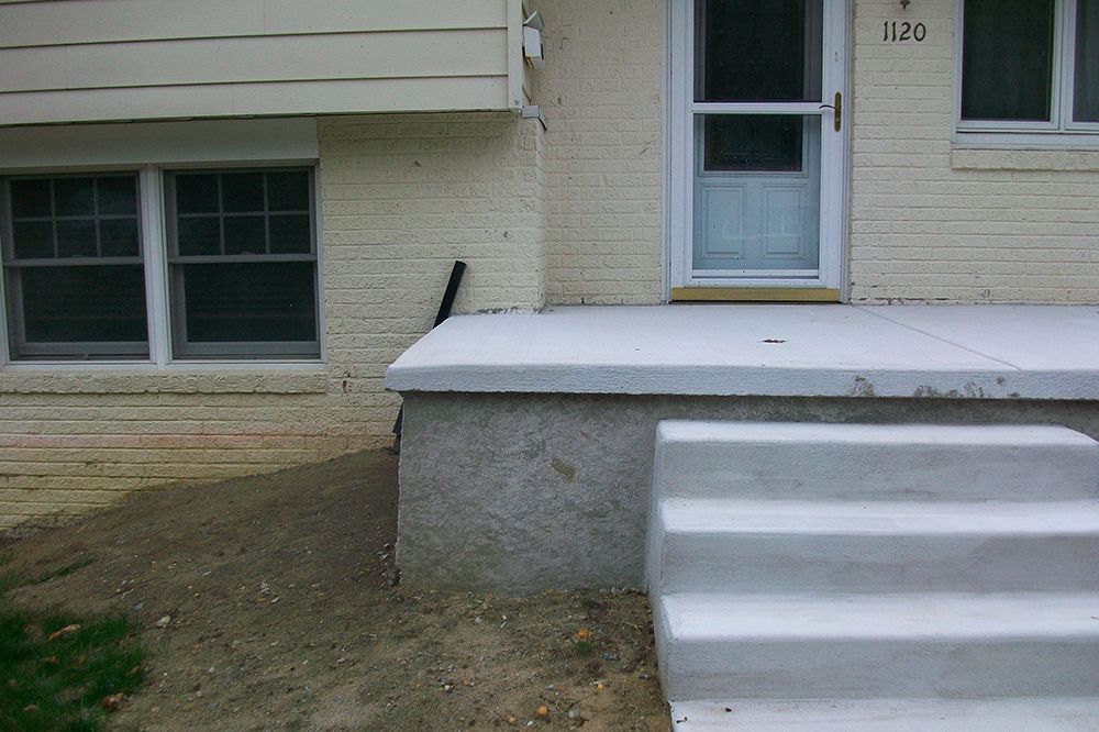 Exterior view of a house with a concrete porch and steps; a window is on the left, and a door is on the right.