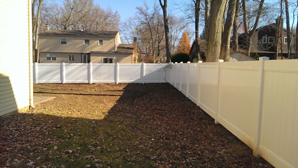 White vinyl fence surrounds a yard with a house and trees in the background. Brown leaves cover the ground.