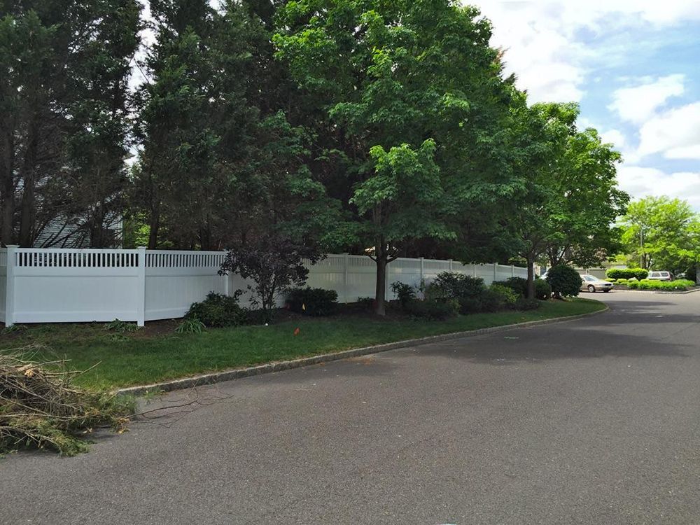 White fence bordering a lush green yard with trees and a paved street.