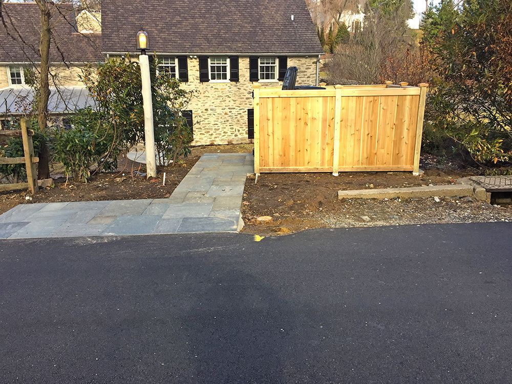 A wooden fence next to a paved driveway leading to a stone house.