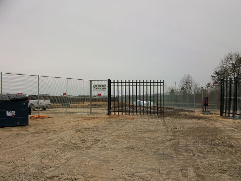 Metal gate in a chain-link fence on a dirt lot. A dumpster and a white truck are nearby under a cloudy sky.